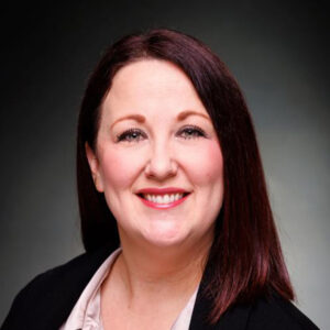 Woman with straight dark hair, dressed in a black blazer, smiling at the camera with a dark background behind her.