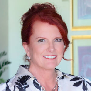 Woman with red hair smiling, dressed in a patterned blouse and necklace, standing indoors with framed artwork in the background.