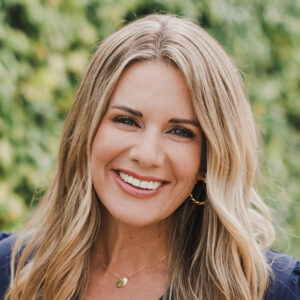 Smiling woman with long blonde hair stands outside in front of greenery, wearing a navy top and gold necklace. Brittany Wermert, RN, BSN, presents a warm and professional demeanor in this natural setting.