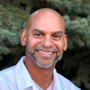 Smiling bald man with a neatly trimmed beard, dressed in a white shirt, standing in front of green pine trees.