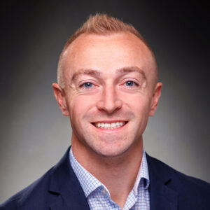 Smiling man wearing a blue suit jacket and checked shirt, standing in front of a dark gradient background.