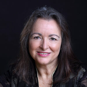 Smiling middle-aged woman with long brown hair, dressed in a black top, standing in front of a dark background.
