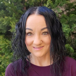 Woman with wavy black hair wearing a maroon top, smiling outside in front of green foliage.
