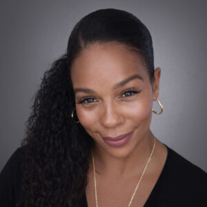 Woman with long curly hair tied in a ponytail, wearing gold hoop earrings and a black top, smiling at the camera.