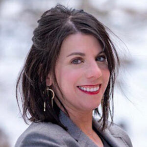 Woman with dark hair in a gray blazer smiling outside, with a blurred, snowy landscape in the background.